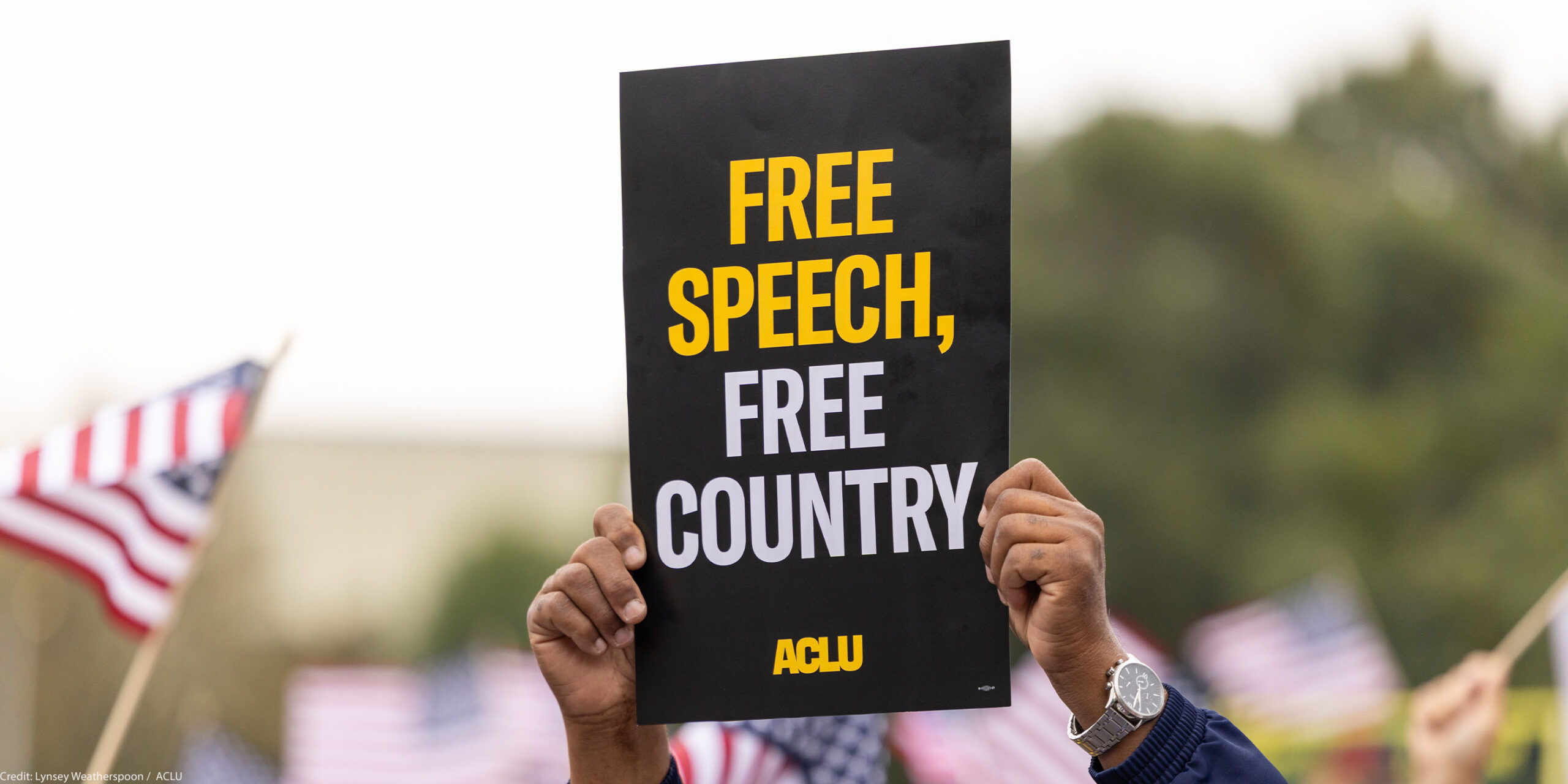 A demonstrator (with only their hands visible) holds up a sign that reads " FREE SPEECH, FREE COUNTRY - ACLU".