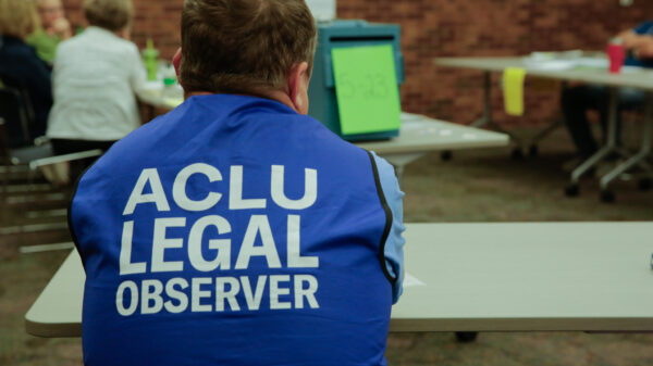 A man sitting at a table at a polling locations wearing a blue vest that reads "ACLU Legal Observer"