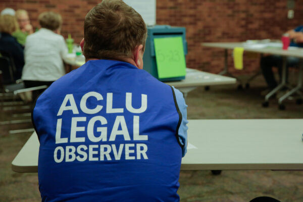 A man sitting at a table at a polling locations wearing a blue vest that reads "ACLU Legal Observer"