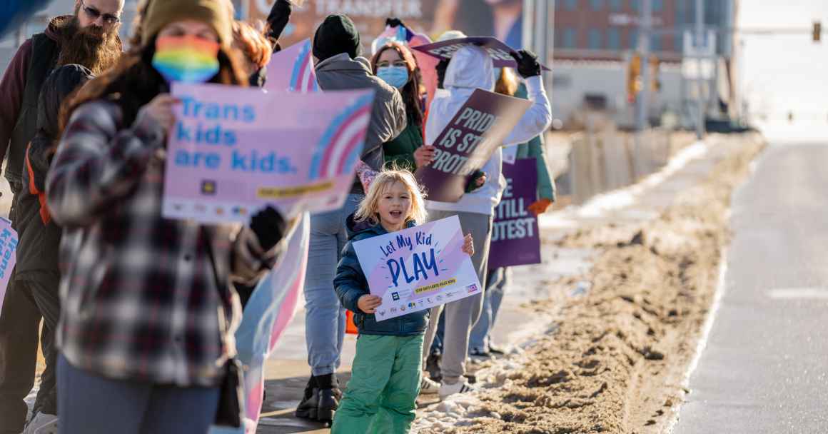 Image of a kid holding a sign that says, "Let trans kids play"